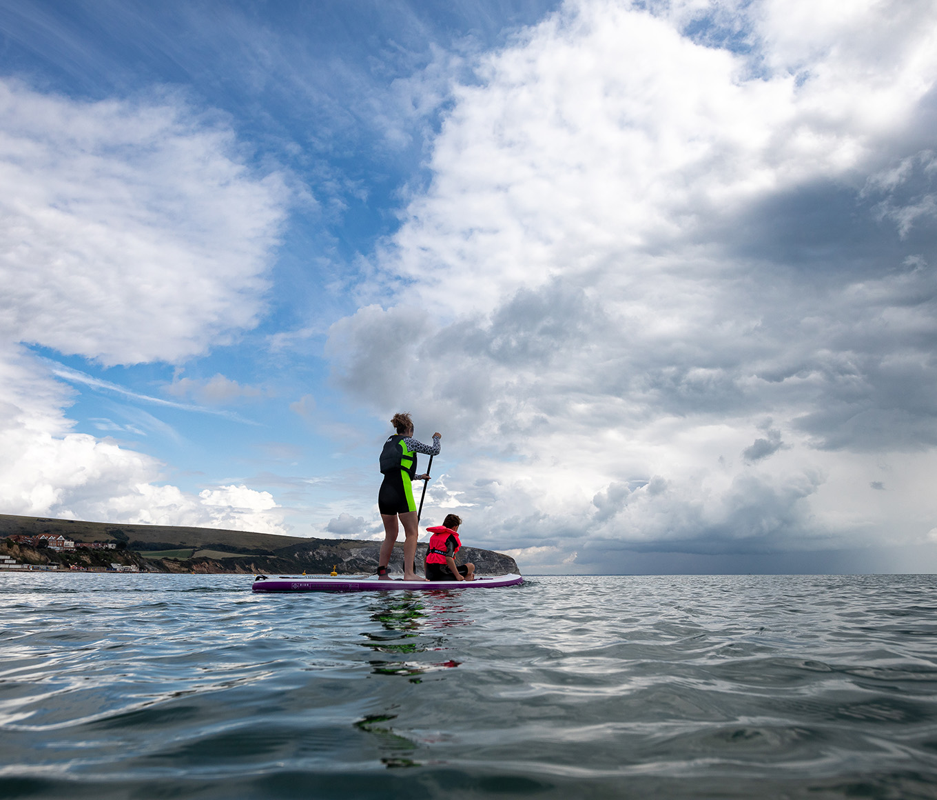 A paddleboarder with a child on the front at Swanage on a bright but cloudy day