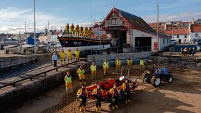Group shot of RNLI volunteers stood outside Anstruther Lifeboat Station with their inshore and all-weather lifeboats and launch vehicles