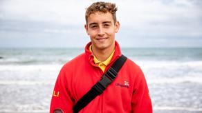 RNLI Lifeguard Hugo is smiling at the camera and is stood in front of the sea on Llangrannog Beach, Wales