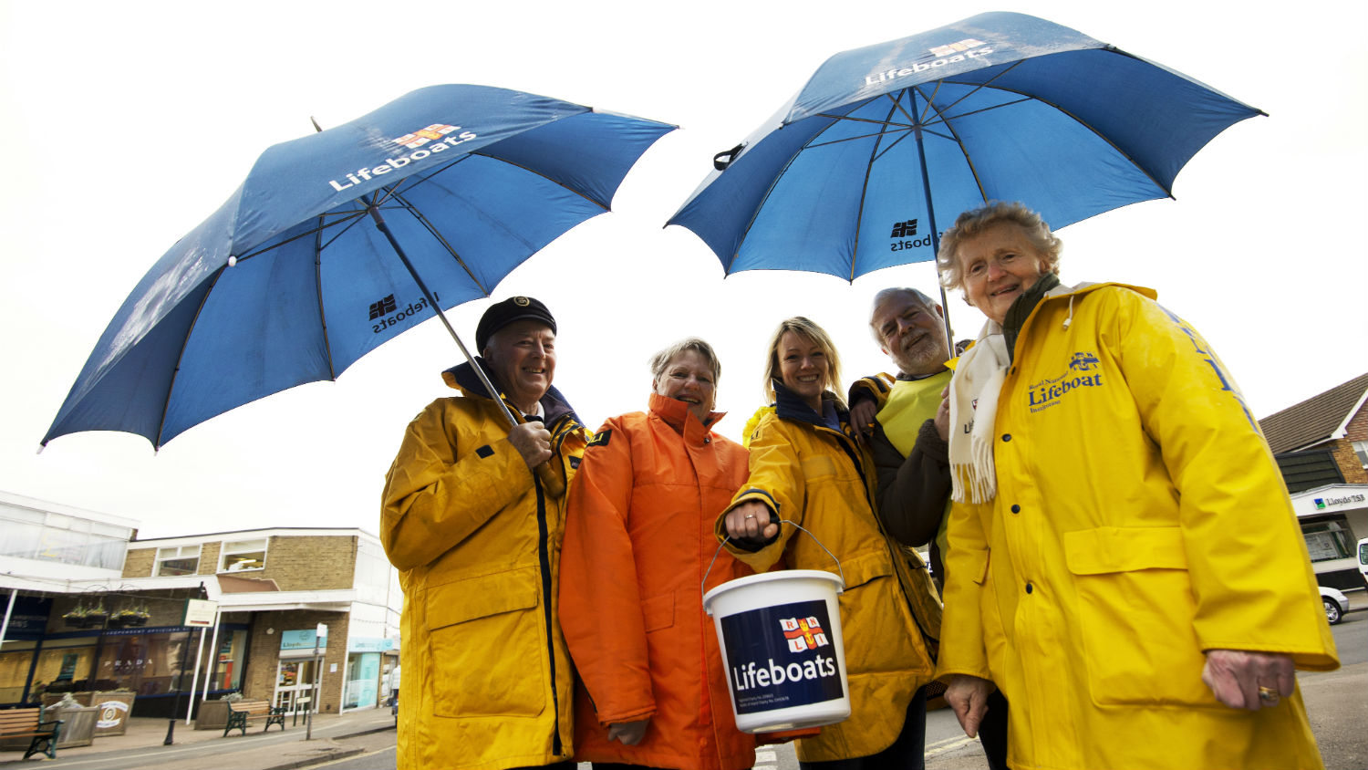 Image of volunteers wearing yellow raincoats stood under umbrellas with a collection bucket