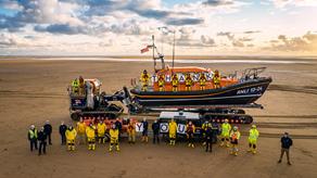 A group of RNLI volunteers standing alongside their lifeboat on a beach on a sunny day. They are holding up cards spelling the word ‘thank you’. 