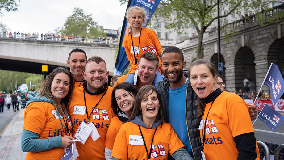 A group of RNLI cheering volunteers at the London Marathon. The volunteers are smiling at the camera and are wearing bright orange RNLI lifeboats T-shirts.