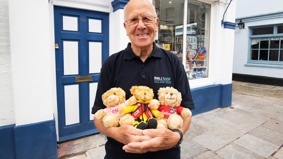 A smiling RNLI shop volunteer standing outside an RNLI shop holding three teddy bears.