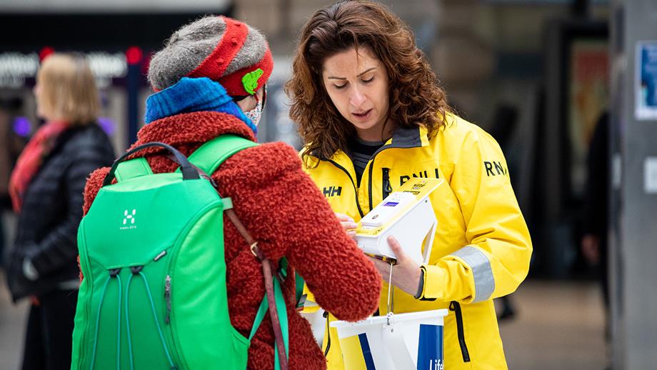 A supporter making a donation with RNLI Fundraising Volunteer and Tower Crew Member Suzanne Goldberg on London Lifeboat Day at Waterloo Train Station. Suzanne is wearing yellow crew kit and is carrying a collection bucket and a contactless payment unit.