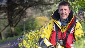 A white lifeboat crew member with short brown hair and a moustache sits in full kit among flowers, with trees in the background