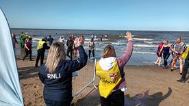 RNLI volunteers on the beach at the Rhyl Winter Dip event. Swimmers are coming out of the water. It’s a bright, sunny day with clear blue skies.