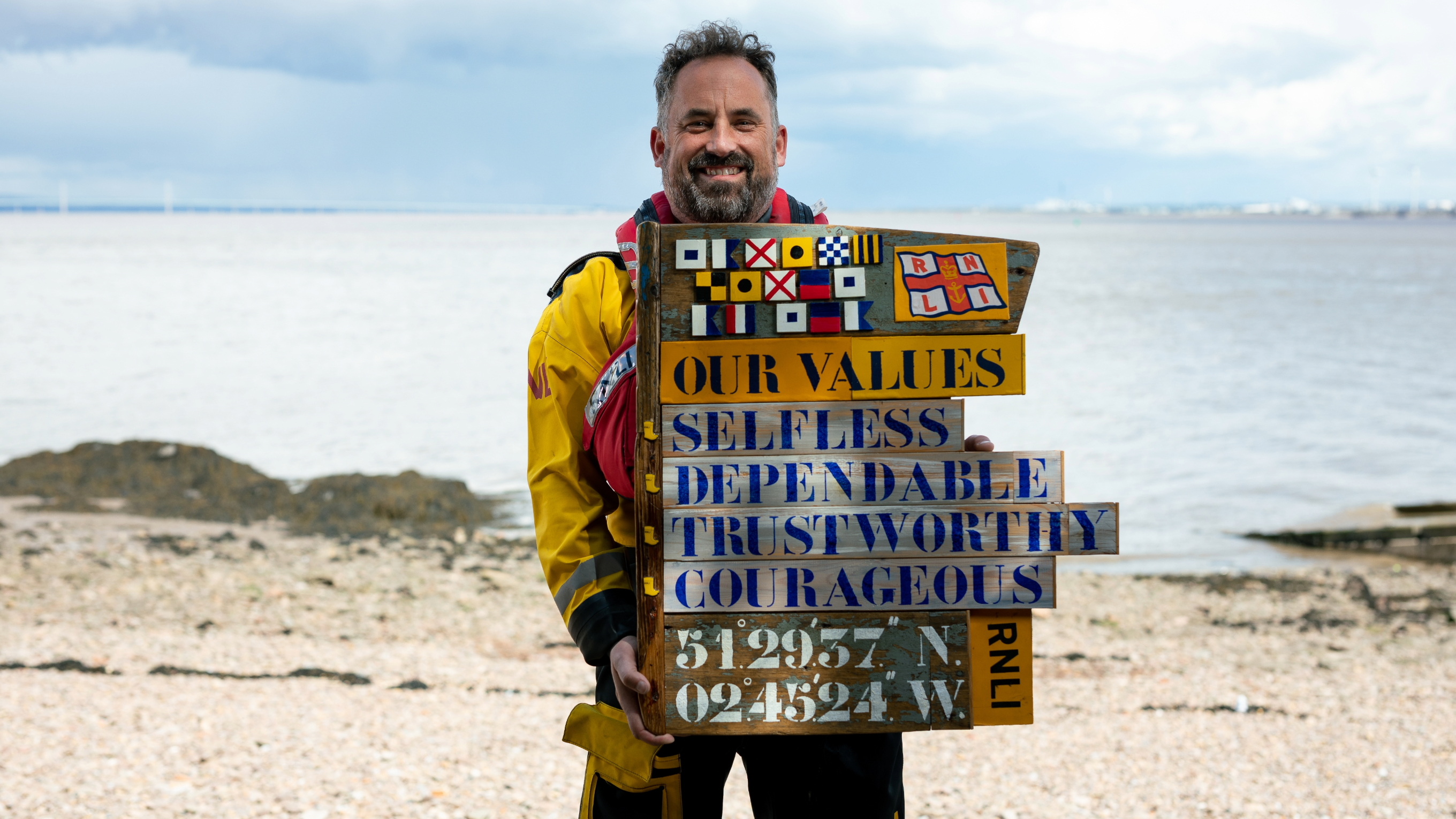 Portishead Lifeboat Station's Helm holding a wooden sign with the four RNLI values printed on it.