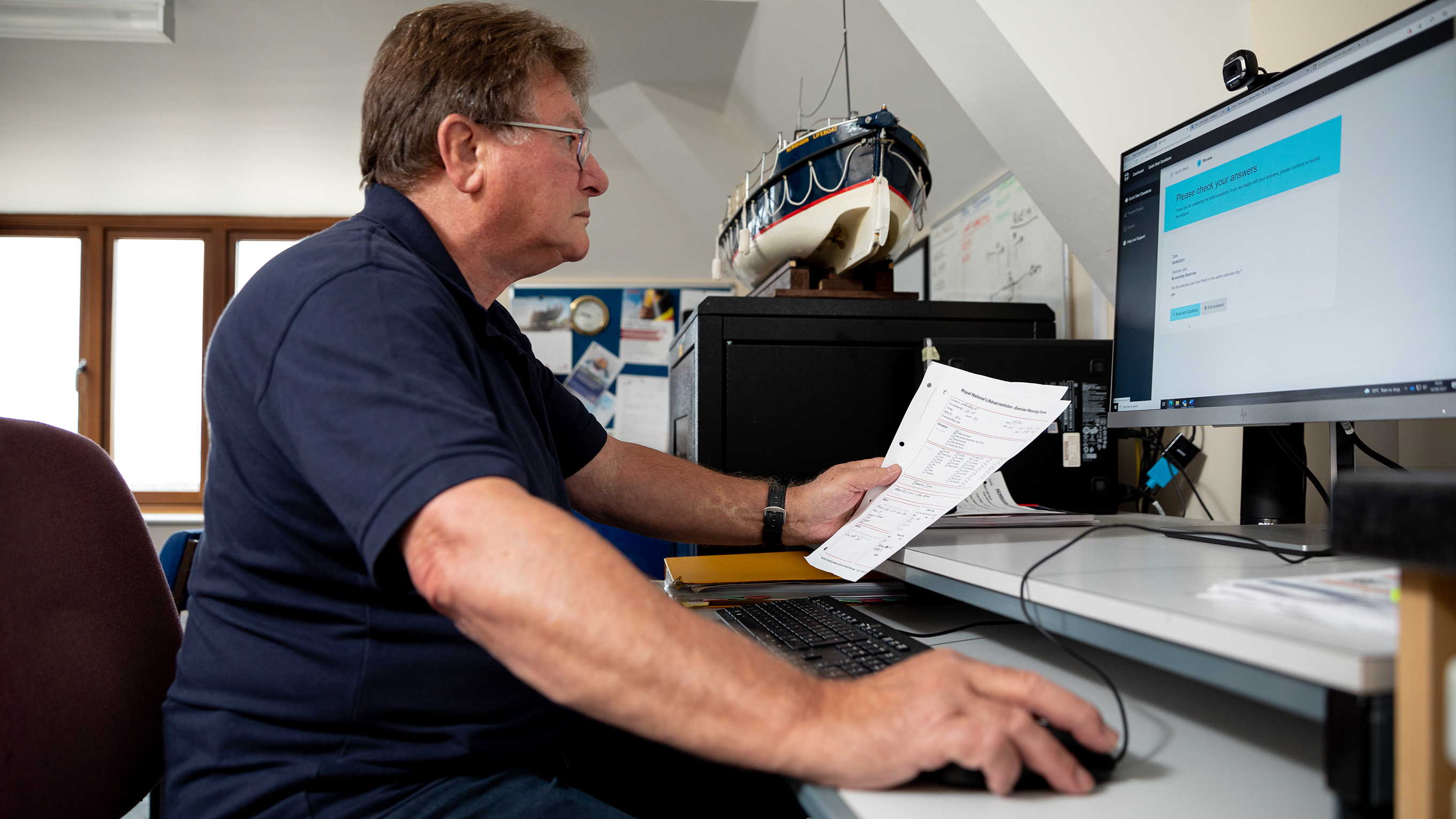 A volunteer working on a computer. There is a model lifeboat on top of a filing cabinet in the background.