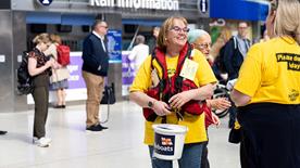 Two fundraising volunteers at a train station holding collection buckets. Members of the public are in the background.