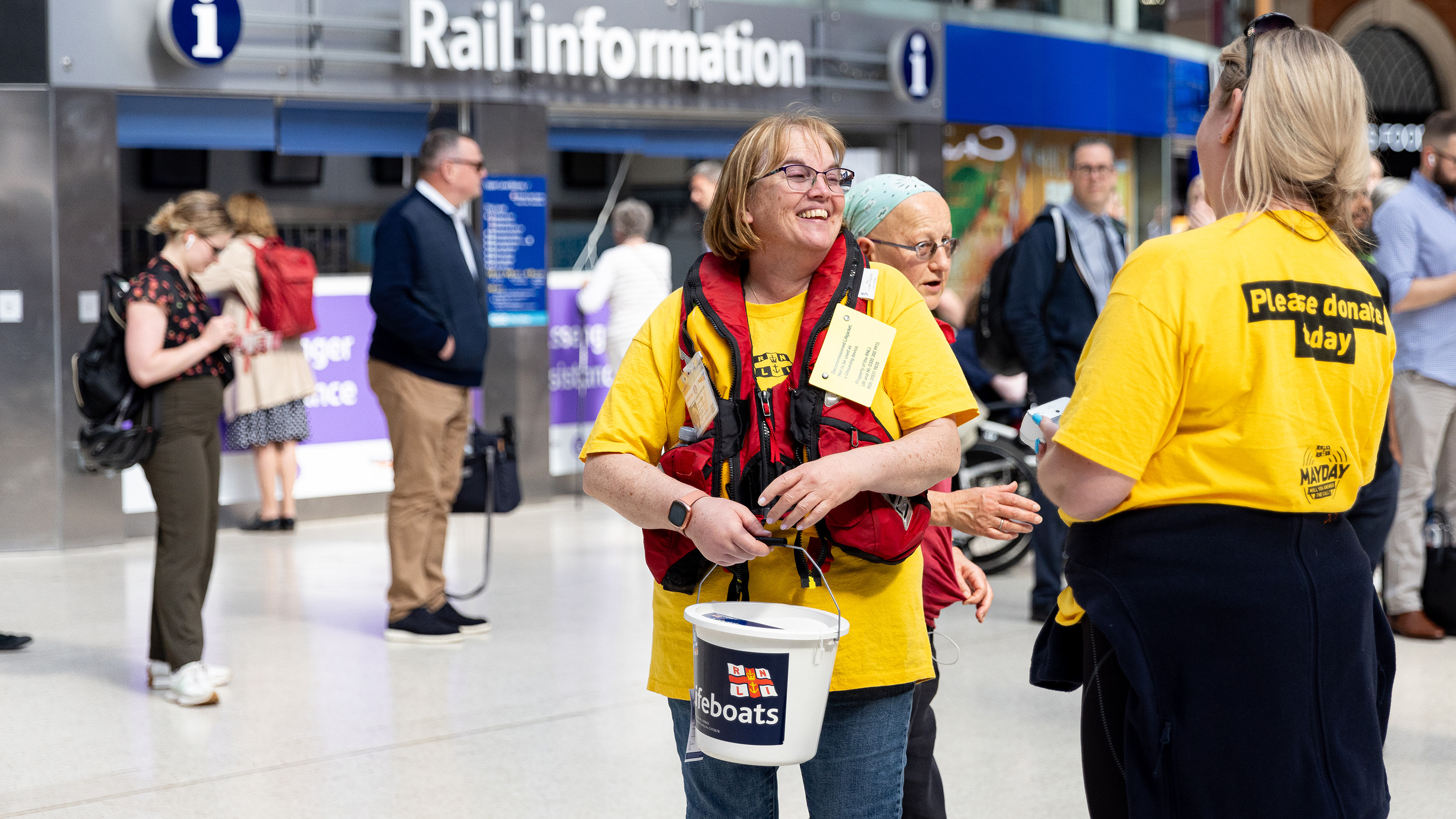 Two fundraising volunteers at a train station holding collection buckets. Members of the public are in the background.