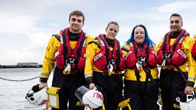 Four volunteer crew members stood in a line wearing their crew kit. Three of them are holding their helmets. The sea is in the background.