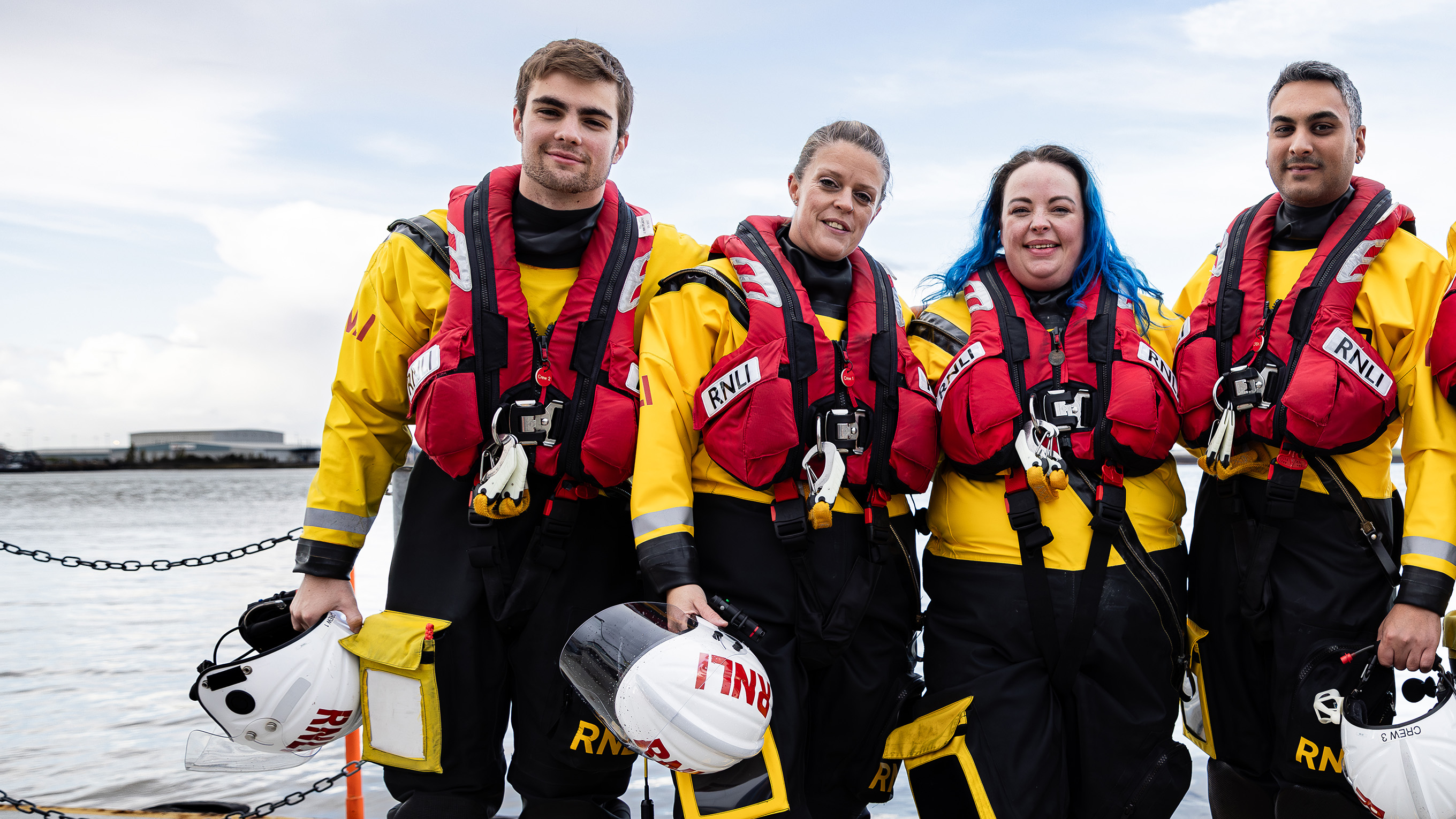 Four volunteer crew members stood in a line wearing their crew kit. Three of them are holding their helmets. The sea is in the background.