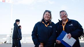 Two volunteers are standing on a beach promenade. They are both smiling and wearing RNLI branded jackets. One of the volunteers is holding a notebook and a clipboard, and the other volunteer is holding a flask. There are two people walking on the promenade in the background.