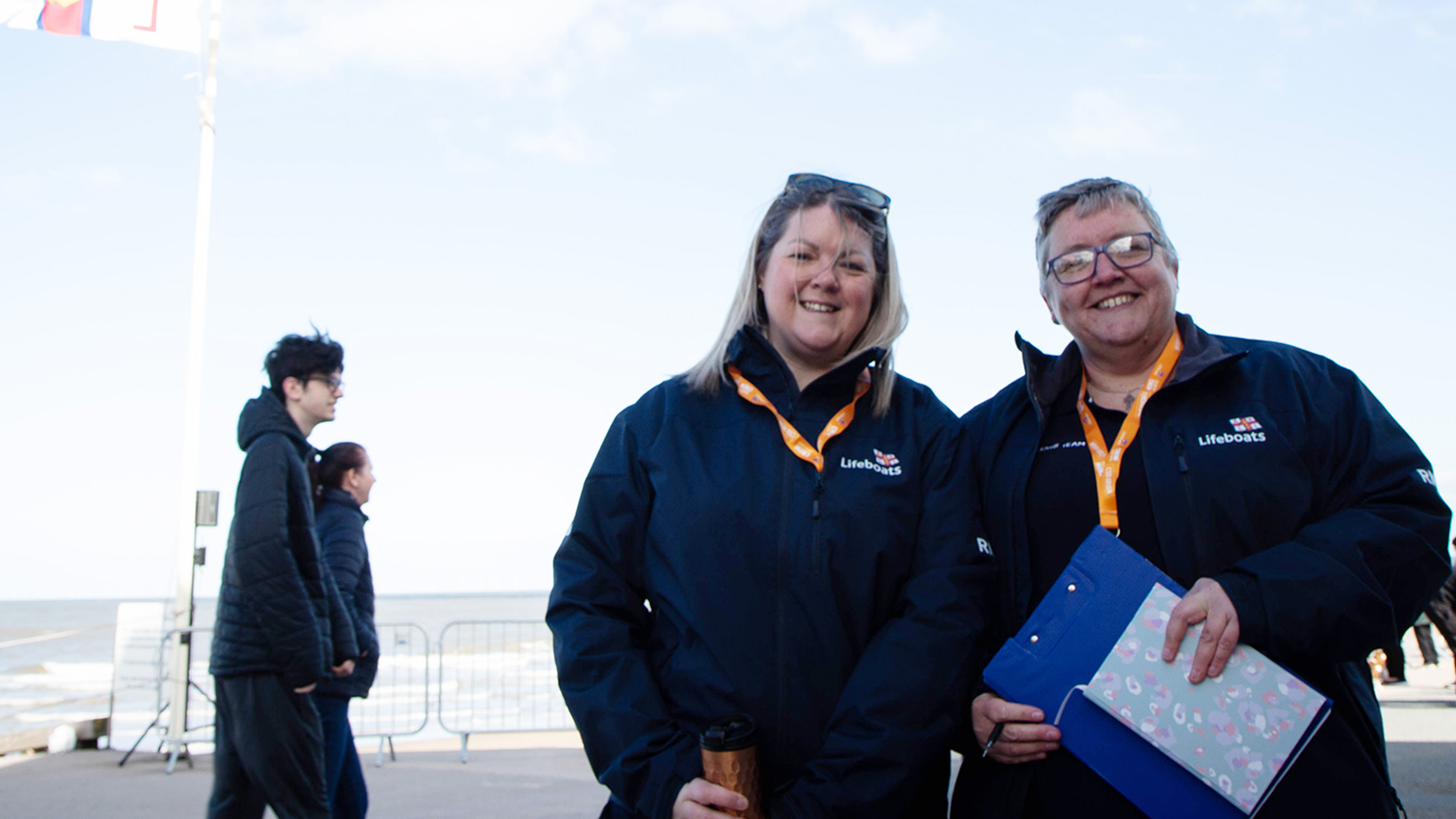 Two volunteers are standing on a beach promenade. They are both smiling and wearing RNLI branded jackets. One of the volunteers is holding a notebook and a clipboard, and the other volunteer is holding a flask. There are two people walking on the promenade in the background.
