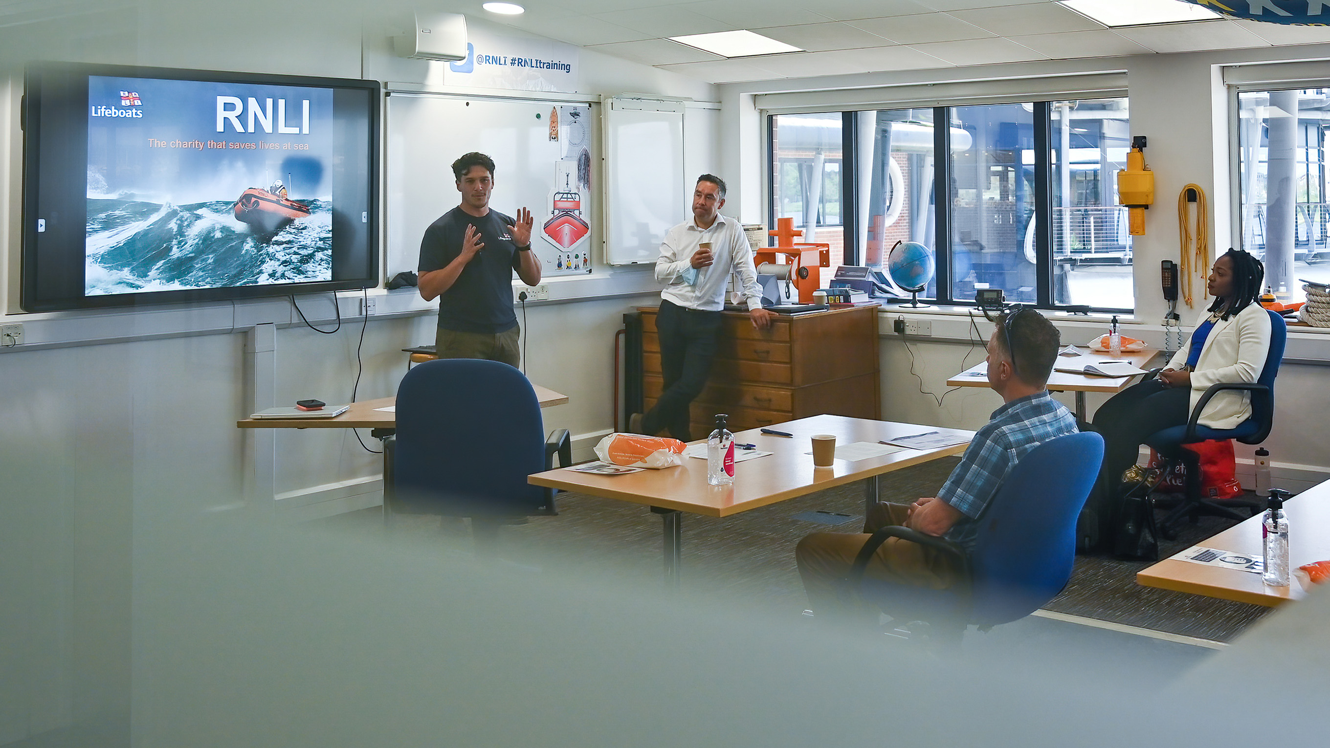 Four people are in a training room at the RNLI College. Two people are sitting at office desks, and another man stands at the side of the room. They are watching a man present some training, and there is an RNLI branded presentation on the screen behind him.