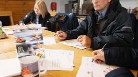 Volunteers gathered round a kitchen table writing things down on paper. On the table, there is a card with lifeboats on, as well as a mug with a lifeboat on.