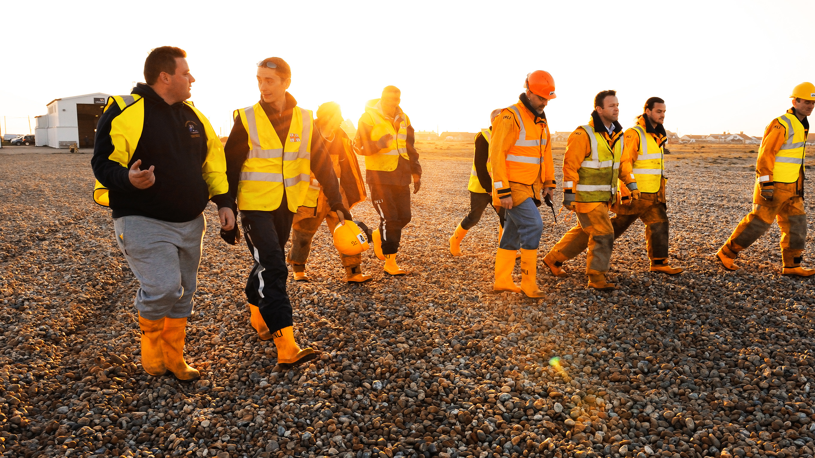 A group of lifeboat station volunteers walking along a pebble beach at sunset.