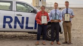 Three people stood on the beach holding awards. There is an RNLI lifeguard vehicle behind them.