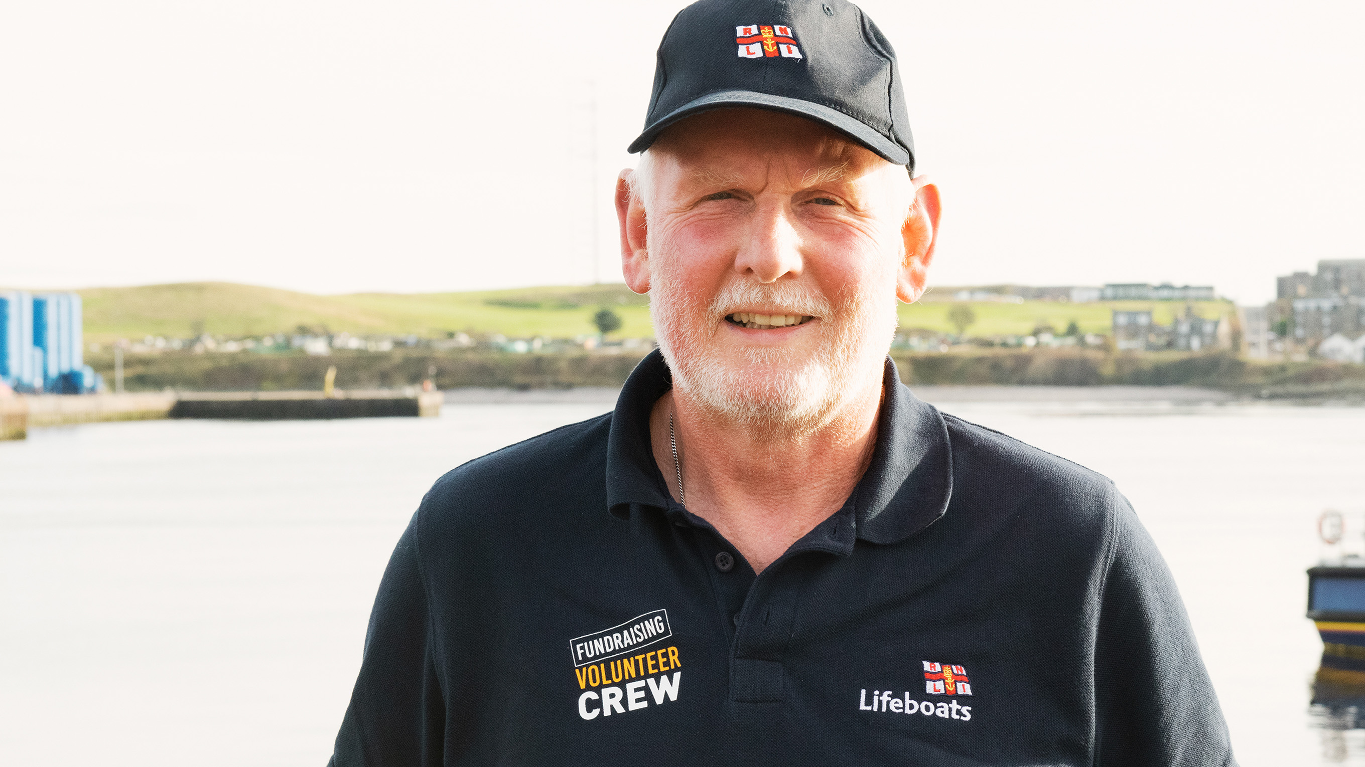 A fundraising volunteer smiling at the camera. He is wearing an RNLI branded polo shirt and cap.