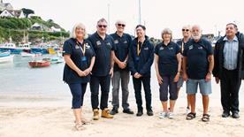A group of volunteers wearing RNLI branded clothing stood on a sandy beach. There are boats on the water in the background.