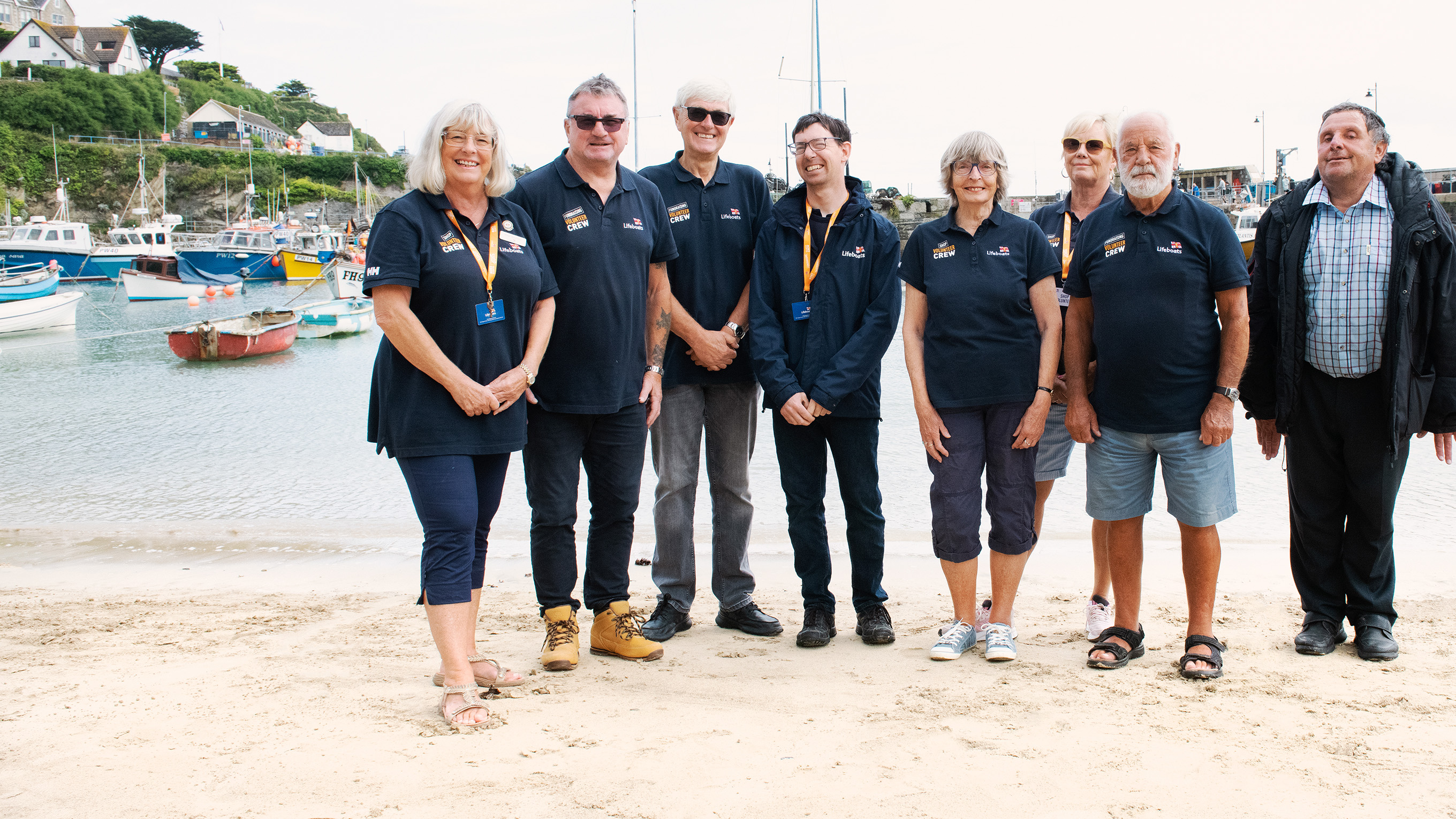 A group of volunteers wearing RNLI branded clothing stood on a sandy beach. There are boats on the water in the background.
