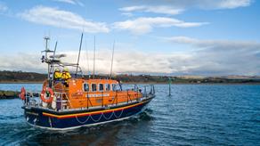 A Mersey class all-weather lifeboat is heading out to sea on a calm, sunny day. Two crew members are standing in the centre of the deck, facing the horizon. 