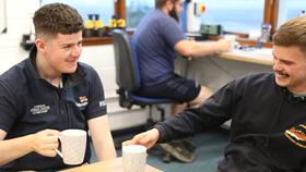 Two RNLI crew members sat at a desk inside a lifeboat stations, smiling and holding mugs of tea