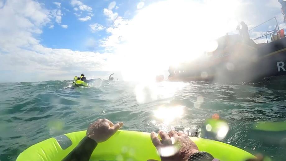 A view from a person in the sea holding onto an inflatable device while looking towards an RNLI lifeboat, with multiple other people in the water