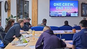 People sat at desks looking towards a screen displaying the words: 'One crew, one you'