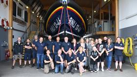 A large group of people standing or kneeling in a row in front of an all-weather lifeboat which is inside a lifeboat station house.