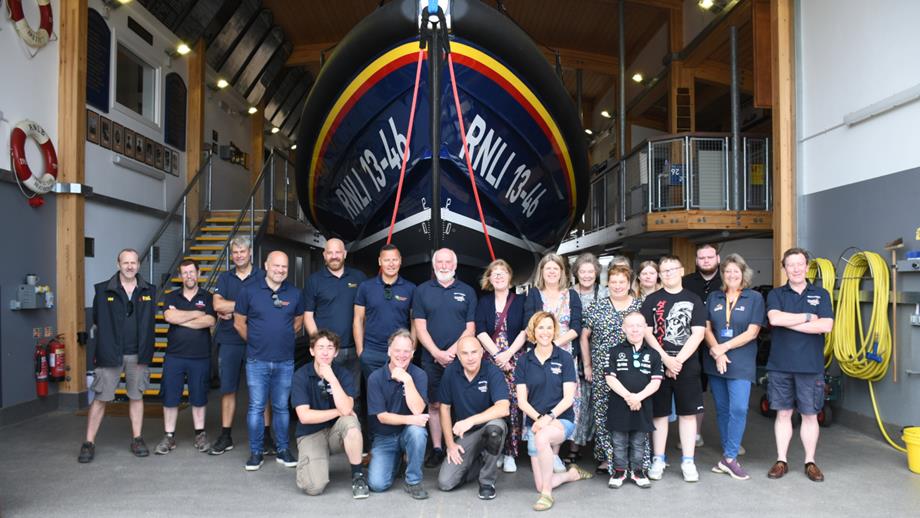 A large group of people standing or kneeling in a row in front of an all-weather lifeboat which is inside a lifeboat station house.