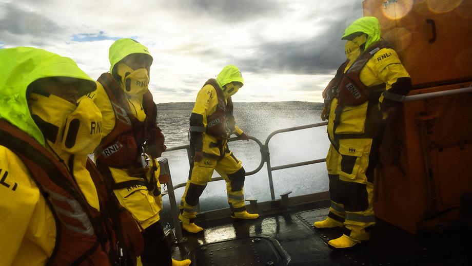 Four lifeboat crew members are out on deck at the stern of an all-weather lifeboat in a rough sea. They are wearing waterproofs, wellies and lifejackets, and they have high-visibility hoods up. They are bracing themselves against the weather.