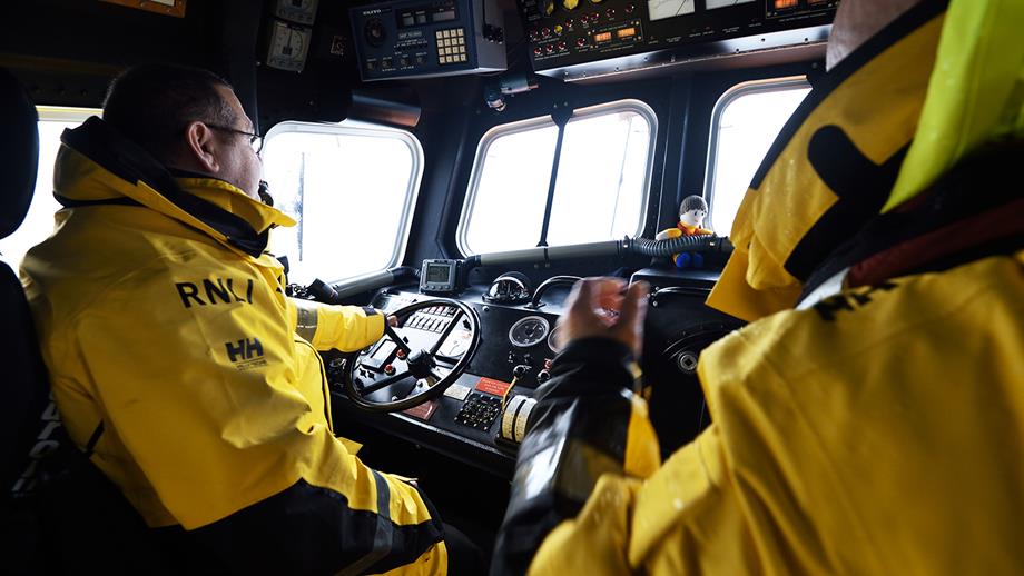 Two crew members are seated inside an all-weather lifeboat, wearing waterproofs. One has his hand on the wheel, steering the boat at sea. There are many controls, buttons and gauges, and windows to the front and to the side of his seat.