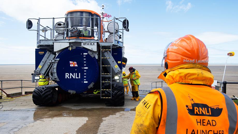 A launch and recovery tractor has brought a lifeboat back to station from the sea, over a wide sandy beach. Three volunteers in waterproofs and high-visibility vest are washing the lifeboat with hoses. One volunteer looks on, with Head Launcher written on the back of their vest.