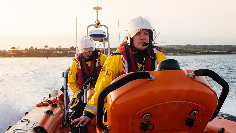 Two crew members are onboard an inshore lifeboat at sea. The sea is calm and the crew members are wearing waterproof kit, lifejackets and helmets. 