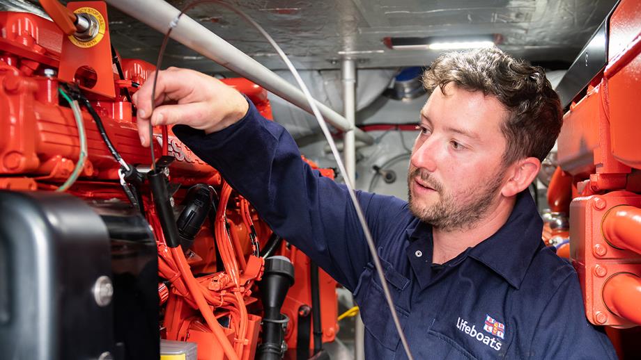 A lifeboat crew member carries out maintenance work on a lifeboat engine. There are two large engines, and the crew member wears mechanic’s overalls.