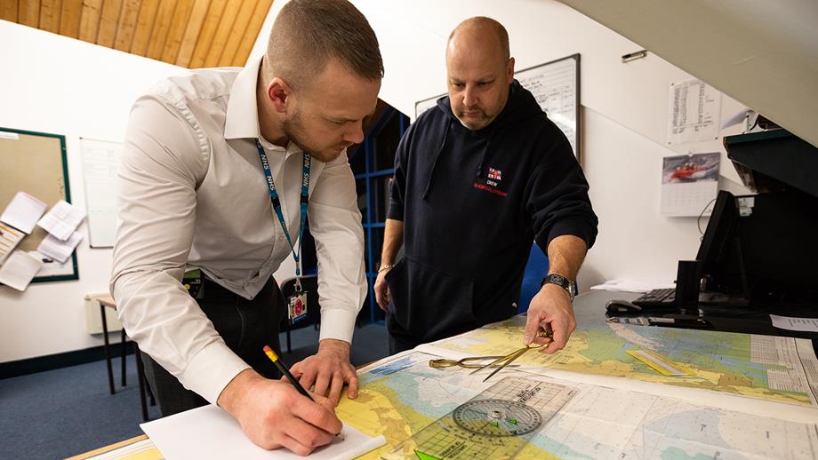 Two lifeboat crew members are doing navigation training in a lifeboat station. There is a maritime chart on a table, and the crew members are using instruments, pencil and paper to practise navigating.
