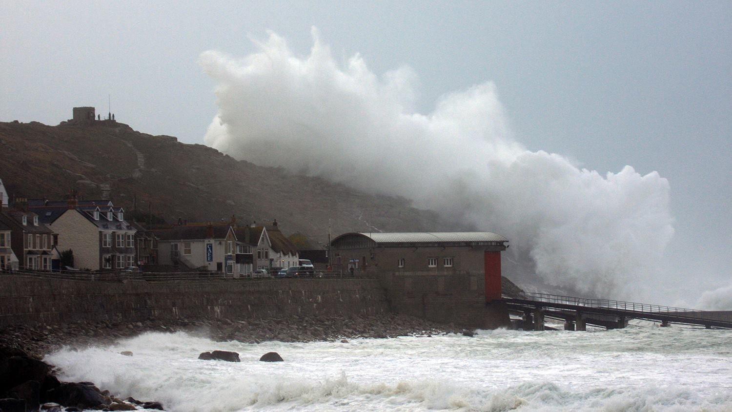 A huge wave breaking over the headland at Sennen Cove during a storm with the lifeboat station in the foreground 