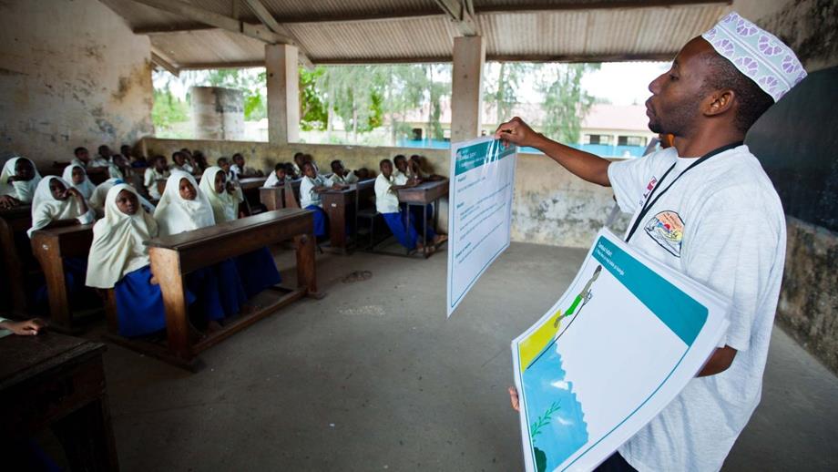 An Aquatic Survival training session taking place in Zanzibar. A teacher dressed in white stands in a busy classroom while showing cue cards with safety messaging.