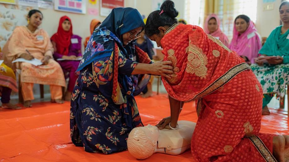 A woman is performing chest compressions on a first aid mannikin in a classroom setting. She is being guided by another woman. In the background, a row of women are seated, watching.