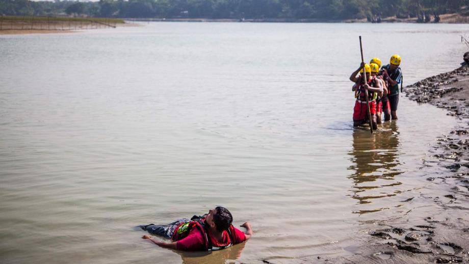 A group of lifesavers in helmets and lifejackets wade through water towards a male casualty in the water as part of a flood safety exercise in Bangladesh.