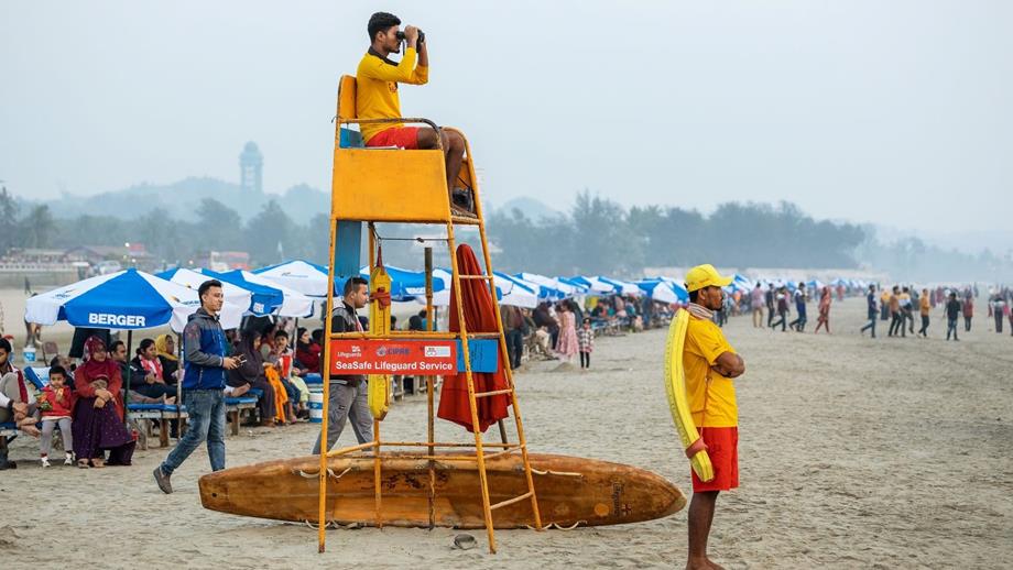 Two male lifeguards are watching over a busy beach in Bangladesh. One lifeguard is sat on a tall seat with binoculars, one is stood on the stand with his arms folded.