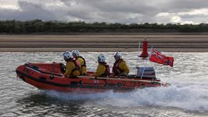 RNLI crew dressed in yellow kit ride a D-Class orange lifeboat fast across a body of water.