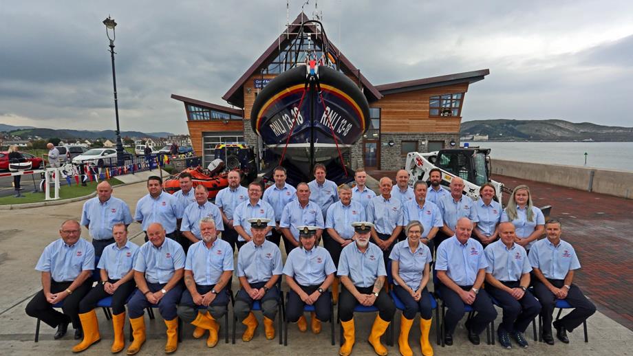 A group of volunteers are standing and sitting in front of the new Shannon class lifeboat and new Llandudno Lifeboat Station building