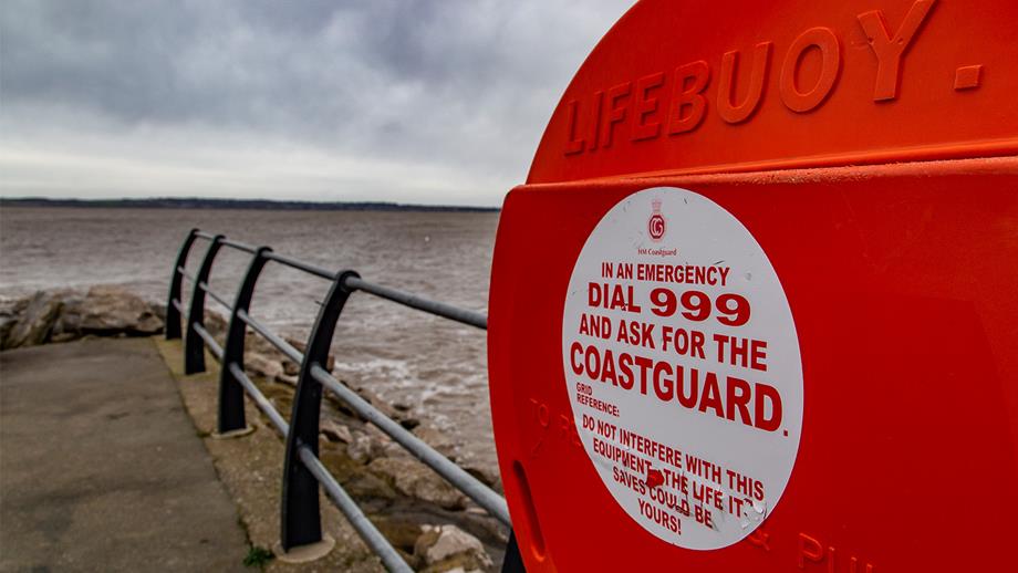 Public rescue equipment at the coast. A view of a circular red lifebuoy casing on a pier with safety railings and the sea in the background. 