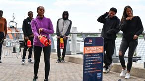 A group of young black adults taking part in water safety and throwbag training outside. There is also a board with details of how to make an emergency call.
