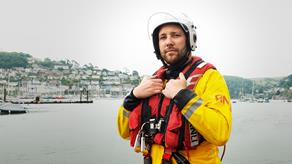 A crew member, wearing  his crew kit and a white helmet, looking directly at the camera. Behind him, you can see the coastal town of Dartmouth.