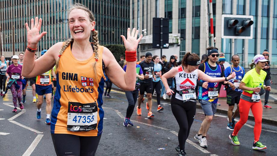 A woman running the marathon for the RNLI smiles at the camera with her hands in the air. There are other runners in the background.