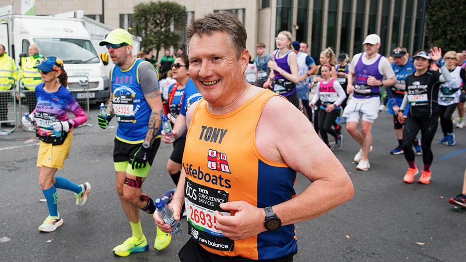 A man smiling as he runs the London Marathon for the RNLI. He is wearing a bright orange RNLI running vest and carrying a water bottle. There are other runners in the background.
