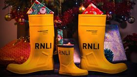 Download this festive RNLI Zoom background featuring two large yellow wellies, beside a junior sized welly, filled with presents underneath a decorated Christmas tree.